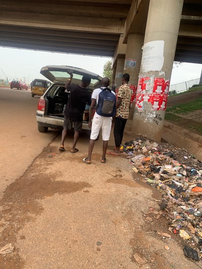 The image depicts a scene under an overpass in Nyanya, Nigeria. Three men are gathered around the open trunk of a car. The man on the left, wearing a black t-shirt and shorts, is looking into the trunk. The man in the middle, wearing a white t-shirt and white shorts, has a blue backpack on and appears to be interacting with the man in the trunk. The man on the right, wearing a patterned shirt and black pants, is standing slightly behind the other two. The trunk of the car is open and appears to contain some equipment. In the background, a large pile of refuse is visible next to a concrete pillar adorned with posters. Some of the visible text on the posters includes "More Than An App" and "SPIRITUAL FATHER". Traffic, including a yellow "keke" (three-wheeled taxi) and a red car, can be seen on the road in the distance. The ground is wet and muddy, suggesting recent rain. The lighting indicates it is daytime, likely overcast.