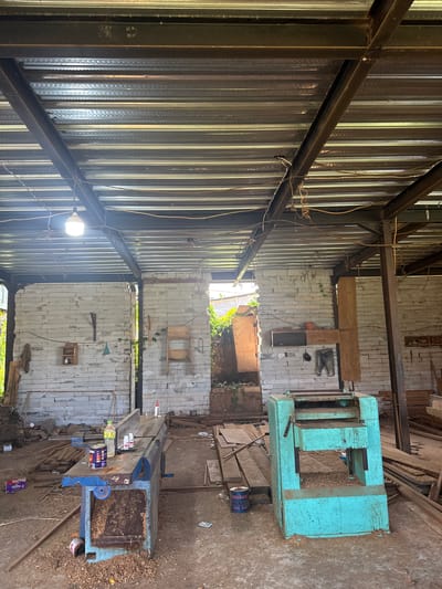 This image depicts the interior of a workshop, likely in Tampaksiring, Indonesia, characterized by its rustic and utilitarian nature. The ceiling is constructed from corrugated metal sheets supported by exposed steel beams, with electrical wiring and a bare lightbulb hanging from above. The walls are made of light-colored, horizontally stacked bricks.
In the foreground, to the left, is a large, blue woodworking machine with a flat, wooden workbench on top. Several cans and bottles of liquids, likely adhesives or finishes, are placed on the workbench. Wood shavings are scattered on and around the machine, indicating recent use. To the right, a bright turquoise industrial planer machine stands, also surrounded by wood shavings.
The floor is made of packed dirt, littered with wood scraps, planks, and various tools. In the background, an opening in the wall reveals an overgrown, outdoor area with lush green vegetation. The overall impression is one of an active but unrefined woodworking space, possibly a small business or individual artisan's workshop.