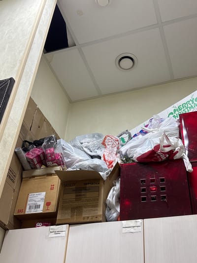 In the city of Ladozhskoe Ozero, Russia, a cluttered storage space is depicted. The image focuses on a dense collection of various items piled high, obscuring much of the room. Dominating the scene are numerous cardboard boxes, some open and others sealed, containing what appears to be merchandise or supplies. Among these boxes are several plastic bags, some filled and tied, others appearing to be empty.

Prominently displayed is a large red cabinet or container, its surface reflective and featuring a grid of nine square, dark openings. Various labels are visible on the boxes, including one that reads "EXPORT GUINOT," and another in Russian that translates to "PERCHATKI" (Gloves) and "VINIL" (Vinyl). Another label indicates "Mastera podtegovogo servisa" (Master of the flooring service) and a sign that reads "Администраторы" (Administrators).

A portion of the ceiling is visible, composed of white acoustic tiles with a ventilation grille. A dark opening is present in the ceiling to the left, suggesting a possible access point or structural anomaly. The overall impression is one of disarray and a significant accumulation of stored goods in a commercial or administrative setting.