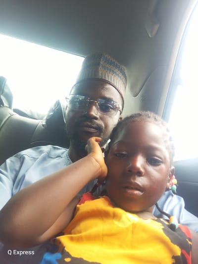 The image is a close-up selfie taken inside a vehicle, likely in Sabonjibi, Nigeria. A man with a beard and glasses is in the background, wearing a blue striped shirt and a patterned cap. He has his hand on his chin, looking towards the camera. In the foreground, a young child, possibly a girl with braided hair adorned with colorful beads, is positioned closer to the camera. She is wearing a yellow top with a black and red pattern. The background shows the interior of a car, including seats and a ceiling. The lighting suggests it is daytime, with bright light coming from outside. A watermark "Q Express" is visible in the lower left corner.