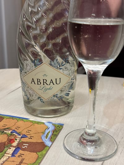A still life image shows a glass of light-colored liquid, likely wine, next to a bottle of Abrau Light Sweet wine. The bottle has a distinctive twisted glass design and a label featuring floral patterns and the text "INTERNATIONAL COLLECTION ABRAU Light SWEET." A corner of a board game with numbered tiles depicting fields, a river, and buildings is visible in the foreground. The scene is set on a light-colored, textured surface, possibly a table. The lighting suggests an indoor setting, potentially a home or restaurant. The image does not provide any information about the city of Kharnavy, Russia, or any individuals present.