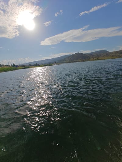 The image depicts a body of water, likely a lake or large river, with rippling dark green water in the foreground reflecting the bright sunlight. The sun is high in the sky, partially obscured by wispy white clouds. In the middle ground, a shoreline is visible with low, rolling hills in the distance under a blue sky with scattered clouds. Along the shoreline, there are signs of human settlement, including what appear to be small buildings, utility poles, and a boat in the water. The scene suggests a natural environment with some human presence. This scene is consistent with locations around Manat, Kazakhstan, known for its natural landscapes and water bodies.