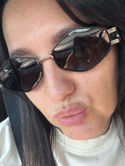 A close-up, selfie-style shot shows the upper half of a woman's face, featuring her eyes, nose, and mouth, partially obscured by sunglasses. The woman has fair skin with freckles visible on her cheeks and nose. Her lips are slightly parted, and she is wearing a light-colored, ribbed sweatshirt. The sunglasses are black and angular with gold accents on the temples. In the reflection of the sunglasses, a smartphone is visible, held by the woman, and a road with cars is seen in the background, suggesting she is in a vehicle. The image does not provide any discernible information about the city of Dol "Luch", Russia.