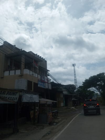 The image displays a street scene in Sukanagalih, Indonesia, under an overcast sky with diffuse lighting. An asphalt road, appearing damp, curves slightly to the right, with a white line marking its left edge. On the immediate left, a series of makeshift commercial stalls are visible, one featuring a sign partially readable as "AR PAINT" and "KILOAN," and another displaying a white banner with a barber pole logo. Behind these stalls stands a multi-story building with a distinctive ribbed roofline and a balcony from which an Indonesian flag (red and white) is suspended. Numerous utility cables are visible crossing the scene above the buildings. Further down the street on the left, additional structures are less clearly defined. On the right side of the road, a dark-colored SUV with illuminated taillights is observed driving away from the camera. In the background on the right, a tall lattice-style telecommunication tower rises above a line of green trees. No discernible individuals or interactions are visible.