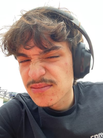 A close-up, upward-angled portrait of a young man with brown, wavy hair, a prominent mustache, and visible facial blemishes on his cheeks and chin. He is wearing black over-ear headphones and a dark-colored crew-neck t-shirt. His eyes are squinted shut, and his nose is wrinkled, indicating a strong facial expression. The background is largely bright white, consistent with an overcast sky or bright diffuse light. A blurry, light-colored, metallic-like structure with an open lattice design is partially visible in the lower-left background. The location is the city of Bron, France.