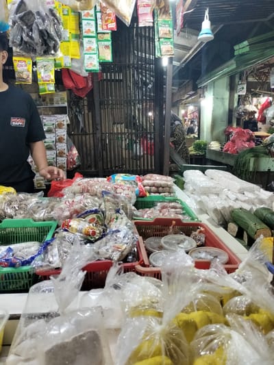 The image depicts a market stall in Bekasi, Indonesia, likely a traditional market, given the open-air setting and the variety of packaged goods. In the foreground, numerous transparent plastic bags are filled with food items, including yellow-colored spices or powdered ingredients, and other unidentifiable food products. Behind these, there are several red and green plastic baskets containing more food items, some of which appear to be meatballs or similar processed foods, also in clear packaging. Further back, various packaged seasonings and snacks are hanging from racks. A person wearing a black t-shirt with a logo that reads "ENGGEM SIAPPP" is partially visible on the left, with their hand extended towards the products. The background reveals more market stalls, with some individuals visible in the distance, suggesting a bustling commercial environment. The overall impression is one of a vibrant local market offering a diverse range of food products for sale.