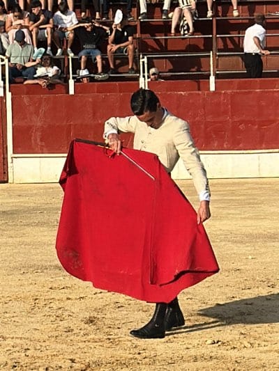 A bullfighter stands in the ring of the Plaza de Toros in San José del Valle, Spain. He is holding a red cape in front of him with both hands. He is wearing a light-colored suit with black boots. The ring is sandy and has a red barrier behind him. There are spectators in the stands behind the barrier.