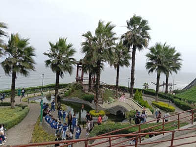 The image shows a park setting in Miraflores, Peru. A pond with a stone bridge spans the water, surrounded by lush greenery, including palm trees. A red-roofed pagoda is visible on a raised platform. Numerous people are present: a group of children and adults, possibly a school group, are clustered near the pond; other individuals are walking along the paths and crossing the bridge. In the background, the ocean meets the horizon. The sky is overcast. A red railing runs along the edge of a path in the foreground.