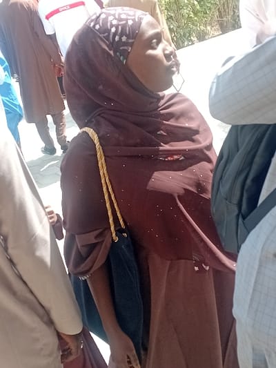 The image depicts an outdoor scene in Kwazailewa, Nigeria, under bright daylight. The central figure is a young adult female, seen in profile, looking upwards and to her right with her mouth slightly open. She wears a full-length maroon or brown garment and a matching hijab with a distinct patterned band featuring white, black, and green elements. Small reflective embellishments are visible on her clothing. She carries a dark blue drawstring bag with a gold braided strap over her left shoulder. Her right hand, partially visible, holds a small dark object.
Surrounding the central figure are several other individuals, mostly partially visible. To her immediate right, the torso and arm of a person wearing a light-colored garment are present. To her left, a person is seen from behind, wearing a white t-shirt with a red horizontal stripe and the word "SPACE" visible, brown trousers, and black shoes. Further back, another individual in a brown traditional garment is partially visible. On the far right, a person wearing a light plaid shirt and a dark backpack is also partially visible. The ground surface is light-colored paving, and the background includes a light-colored wall or structure and sparse green foliage.