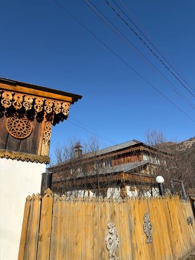 The image presents a low-angle view of buildings in Manzherok, Russia, against a clear blue sky. On the left, a section of a wooden building features intricate carvings, including a decorative rosette. A set of power lines runs parallel to the left edge of the frame. To the right, another building exhibits rustic wooden siding and additional ornate carvings along its edge. Bare tree branches intermingle with the buildings, suggesting it may be winter. A white, cylindrical light fixture is visible on the side of the right-hand building. The camera is angled upward, emphasizing the height of the structures.
