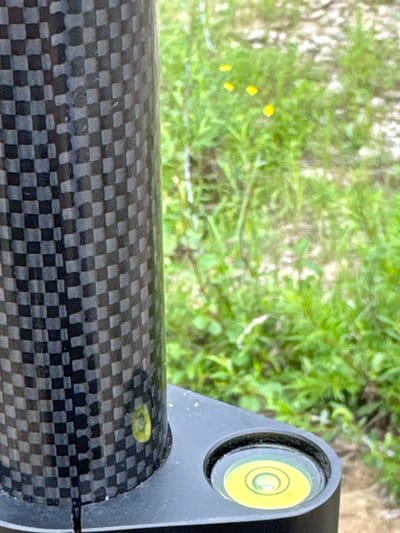 This close-up, eye-level shot focuses on a carbon fiber tube, likely part of a piece of equipment such as a camera tripod or a surveying tool. The tube has a distinctive checkered pattern of black and grey squares. To the right of the tube, a silver bracket is visible, housing a circular spirit level with a yellow bubble. The bubble is positioned slightly off-center, indicating the surface is not perfectly level. In the background, out of focus, is a lush green environment with scattered small yellow flowers, suggesting an outdoor setting in a natural landscape. The lighting appears to be natural daylight, possibly mid-morning or afternoon, and the weather seems pleasant and clear. The scene evokes a sense of fieldwork or outdoor activity, possibly related to photography, construction, or scientific measurement. No text is visible in the image. The location is identified as Murtygit, Russia.
