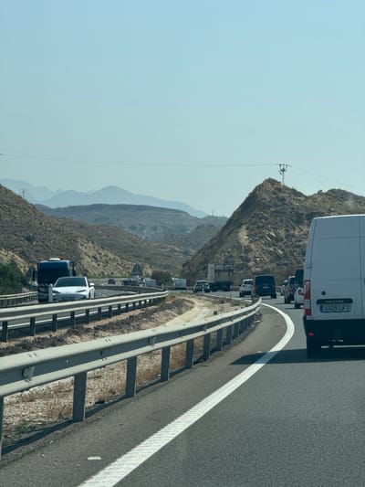 The image shows a multi-lane highway in a mountainous, arid region under a clear blue sky. Traffic is moderate to heavy, with vehicles including cars, vans, and trucks visible in both lanes. The road is paved with asphalt and marked with white lines. A metal guardrail separates the lanes and the roadside. The landscape is characterized by dry, rocky hills covered in sparse vegetation. A white van with the license plate "3329 LFY" is in the foreground, on the right side of the image. The overall scene suggests a sunny day on a highway in a rural or semi-mountainous area of Spain, likely during daylight hours due to the bright sunlight and clear sky.