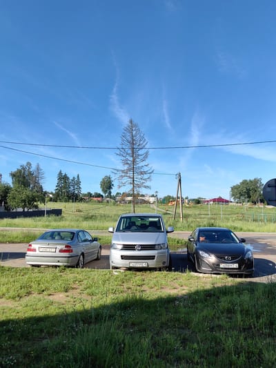 The image depicts an outdoor scene under a clear blue sky with wispy white clouds. In the foreground, three cars are parked on an asphalt surface next to a grassy area. From left to right, the cars are a silver sedan, a silver minivan, and a dark gray coupe. The silver sedan has a license plate that reads "2080 AX-5". The silver minivan has a license plate that reads "4377 PE-5". The dark gray coupe has a license plate that reads "BY 4058 HT-4".
Behind the cars, there is a large grassy field dotted with scattered trees. In the middle ground, a tall, bare, dead tree stands prominently. Further back, a playground area with colorful equipment is visible. Beyond the playground, there are buildings with red roofs, and a residential building with white walls. Power lines and a wooden utility pole are present in the scene. The overall environment suggests a park or recreational area within a town. The license plates indicate the location is likely Belarus, specifically Fanipal, Belarus, as suggested by the context provided.