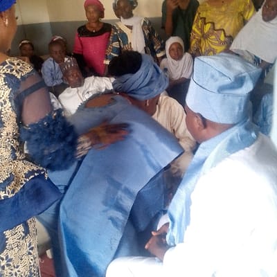 An indoor scene in Ozuri, Nigeria, depicts a gathering of approximately ten to fifteen individuals, predominantly adults with some children, within a room featuring light-colored walls. In the immediate foreground, to the left, a person is partially visible from the back, wearing a dark blue garment with gold embroidery and a textured sleeve. Their right hand, showing traces of henna, is extended forward. To the right foreground, another individual is seen from the back-right, dressed in a white tunic and a light blue traditional cap (Fila), with their hands clasped.
At the center of the image, two individuals are engaged in an interaction. One person, with their back largely to the camera, wears a light blue headwrap and matching traditional attire, extending their left arm in what appears to be an embrace or supportive gesture towards another individual partially obscured opposite them. To the left of this central pair, an older man with a grey beard and a white traditional cap is seated, looking towards the camera. To the right, a young girl in a white hijab is also seated, observing the scene. The background contains several women standing, clad in diverse colorful Nigerian traditional garments and headwraps, alongside other adults and children seated or standing, observing the central activity.