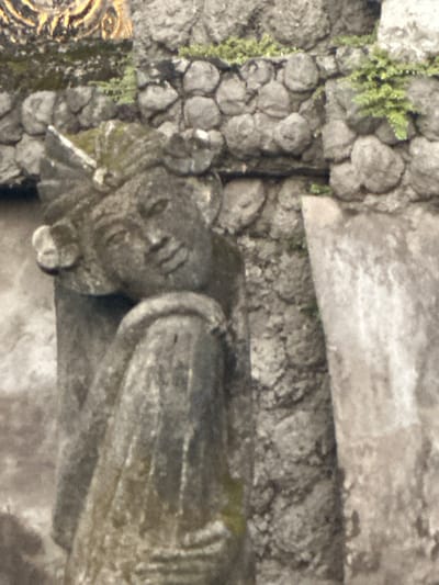 The scene is located in Celuk, Indonesia. The image presents a close-up view of a weathered, dark grey stone statue against a textured stone wall. The statue, depicting the upper torso and head of a figure, occupies the central and left-mid frame. Its head is tilted slightly to its right, featuring a serene facial expression, a headdress with ornate elements, and distinct ear adornments. The statue's surface shows significant weathering, pitting, and patches of green moss or lichen. Behind the statue is a rough-hewn wall constructed from irregularly shaped grey stones, also exhibiting weathering and small growths of green foliage. In the upper left corner, a small, out-of-focus section of an ornate, gold-colored carving is partially visible above the stone wall. To the right of the statue, a lighter, vertically oriented stone slab or column is present. No human actions or interactions are depicted; the scene is static, portraying architectural and sculptural elements within an outdoor environment.