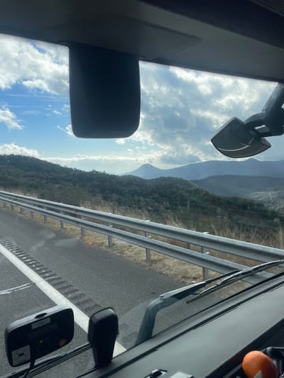 The image depicts a daytime scene from the interior of a vehicle, looking forward through the windshield, presumably within the vicinity of Arbúcies, Spain. The foreground shows internal vehicle components: a black rearview mirror casing centrally positioned at the top edge of the windshield, and a mounting bracket or arm extending from the top right. In the bottom left, a black electronic device, possibly a toll transponder or GPS unit, displaying "99.0" and a green indicator light, is affixed to the windshield. The bottom right reveals a section of the dashboard and two windshield wipers at rest. An orange-colored object, consistent with a fruit, is visible on the dashboard.
The midground features a paved road with a single visible lane defined by a solid white line and rumble strips on its left edge. A double-bar metal guardrail runs parallel to the road on the right, separating it from a dry, light-brown grassy slope. A wire fence is visible behind the guardrail and grass.
The background presents a mountainous landscape under a partly cloudy sky. Closest to the viewer are hills densely covered with dark green trees and shrubs. Beyond these, multiple layers of progressively lighter blue-grey mountains recede into the distance. The sky above is a mix of blue patches and scattered white and grey clouds, suggesting an overcast but bright day. No people are visible. The scene implies a vehicle in transit along a highway or main road through a rural or semi-rural mountainous region.