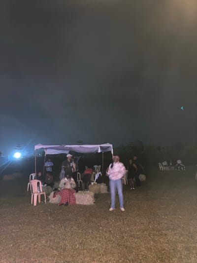 The image depicts an outdoor nighttime gathering in a grassy area within Murera ward, Kenya. In the immediate foreground, center-right, a woman with dark hair stands facing slightly right, wearing a light-colored patterned long-sleeved top, light blue jeans, and white shoes. Her arms are slightly bent at the elbows.

In the midground, a white canopy structure, supported by poles, serves as a central point. Underneath and around this canopy, approximately 8-10 individuals are gathered; some are standing, while others are seated on white plastic chairs. Several rectangular straw bales are positioned around the canopy, with a red and black plaid fabric draped over one. One individual under the canopy wears a light-colored hat.

To the far left of the canopy, a bright, circular artificial light source is visible. In the background, to the right, a separate cluster of white plastic chairs and at least two white tables are arranged, with approximately 3-4 individuals seated around them. The ground consists of short, dark grass, and the sky above is dark, indicating a nighttime setting. No specific actions of direct interaction are clearly discernible, but individuals appear to be congregated in groups.