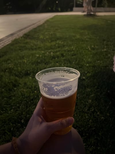 A person's hand is holding a clear plastic cup filled with amber-colored liquid, likely beer, with a frothy white head, in a dimly lit outdoor setting. The background consists of a green lawn with some scattered white speckles, suggesting it's nighttime. A paved walkway with textured edges runs diagonally across the upper left portion of the image. In the far background, out of focus, are structures that appear to be part of an urban environment, possibly a park or recreational area. The overall lighting suggests it is evening or night, and the focus is on the hand and the drink.