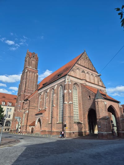 The image shows the St. Martin's Church in Landshut, Germany, a Gothic brick church with a tall, slender tower and a red tiled roof. The church is made of red brick and features arched windows and entrances. Two people are walking on the cobblestone street in front of the church. The sky is blue with scattered white clouds. The scene appears to be a sunny day.