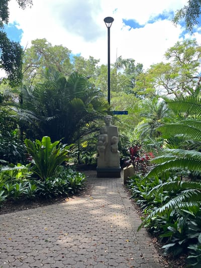 A paved pathway, made of hexagonal interlocking pavers, winds through a lush, tropical garden or park under a bright daytime sky. On the left side of the path, a light-colored stone sculpture depicts a seated figure, likely a woman, with several smaller figures (appearing to be children) gathered around her, possibly engaged in reading from an open book. The sculpture rests on a dark, rectangular base.

To the left of the pathway and behind the sculpture, a tall, dark lamppost with a circular light fixture extends upwards against the sky. Near the sculpture, a blue directional sign is mounted on a pole, displaying a white arrow pointing to the right and an unreadable white icon or text below it.

The garden is densely planted with a variety of green foliage, including large ferns, palm fronds, and other broad-leafed plants, creating a verdant and shaded environment. The sun casts dappled light and shadows across the pathway, suggesting either morning or afternoon. The sky in the background is mostly clear with some scattered white clouds. The tropical vegetation aligns with the provided context of Brisbane City, Australia. No people are actively present, only the figures in the sculpture.