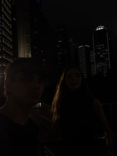 The image is a dark, low-light selfie taken at night, with the camera angled upwards. Two people are visible, a woman in the foreground and a man slightly below and to the left. The woman is looking towards the camera, her face partially illuminated by ambient light. Her hair is long and blonde. The man's face is also visible, with his head tilted upwards.
The background features illuminated skyscrapers, characteristic of a modern city skyline at night. These buildings are tall and imposing, with many windows lit up. One building in the upper left has visible text, appearing to be Chinese characters and possibly a logo or name. Another building to the left of center has vertical lines of light and what looks like a business name written horizontally in Chinese characters. The reflection of the city lights is visible on a wet surface, suggesting it may have recently rained or the area is damp.
The overall mood is intimate and possibly romantic, given the selfie format and the nighttime urban setting. The dark environment and the way the subjects are positioned suggest a close bond between them. The time of day is clearly night, and the presence of rain or moisture in the air adds to the atmospheric quality of the image. The location context of Pudong, China, is consistent with the high-rise cityscape depicted.