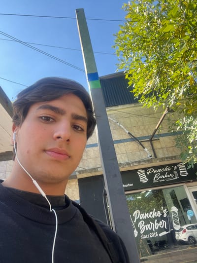 A selfie-style photograph of a young man, presumably in his late teens or early twenties, is taken outdoors. He is wearing an earbud. Behind him is a barber shop called "Pancho's Barber since 2018." The image appears to be taken in Gerli, Argentina, due to the sign and architectural style. The background includes a blue sky, trees, and the facade of the building housing the barber shop, including a window with a partial view of a white car inside.