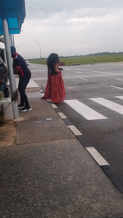 The image shows a paved outdoor area with a walkway and a road. In the foreground, a person wearing a dark top and a long, red patterned skirt stands on the walkway, facing away from the camera and holding a black object. To the left, another person is partially visible, wearing a blue and red polo shirt, dark pants, and white sneakers. This person is leaning forward and appears to be interacting with someone off-camera. The walkway has a concrete surface with a black and white striped curb. Beyond the curb is a wet asphalt road with white pedestrian crossing lines. In the background, a white ambulance is visible on the road, and a cloudy sky stretches above. The environment appears to be an open area, possibly near an airport or a large intersection, with green fields and distant trees. The location is identified as Orerokpe, Nigeria.