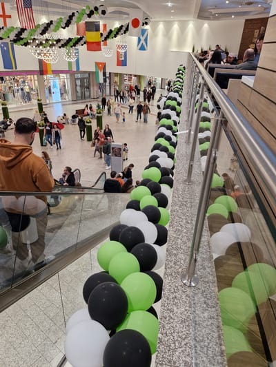 This is an aerial photograph of a shopping mall, possibly in Bremen, Germany, decorated for an event. The mall features multiple levels and an atrium with a tiled floor. Decorative balloons in black, white, and lime green are arranged in a long, curving line along a railing, ascending towards the upper levels. Several national flags, including the US flag, are hung from the ceiling. People are seen shopping, walking, and sitting on benches on the lower level. An escalator is visible in the foreground.