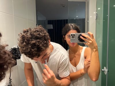 The image displays a mirror selfie of two individuals, one male and one female, in a modern bathroom located in Orléans, France. The male, positioned to the left within the reflection, has dark, curly hair and wears a white t-shirt. He is actively brushing his teeth with a blue toothbrush, his head bent forward. To his left, a wall-mounted black and silver hairdryer with a coiled cord is affixed to the white-tiled wall. The female, positioned to the right and slightly behind the male, has dark hair styled away from her face and wears a white tank top. She wears a necklace with small beads and a central reddish-brown pendant, along with a thin bracelet on her left wrist. She holds a smartphone with a clear case and a black circular grip in her right hand, capturing the reflection. The bathroom walls consist of white square tiles to the left and green rectangular tiles to the right, where a chrome circular shower control is visible. The reflection in the upper portion of the mirror reveals a dimly lit room behind the subjects, featuring vertically striped curtains, a lamp with a white shade, and other elements indicative of an adjacent bedroom or hotel room.