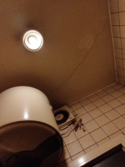 The image shows a bathroom in Riyadh, Saudi Arabia, located at coordinates 24.712, 46.696. A white ceiling fan is mounted on the ceiling. The ceiling is light brown with a crack running diagonally across it. A white water heater is sitting on the floor in the lower left corner. A white electric fan is sitting on the floor to the right of the water heater. The floor is covered in beige square tiles. A small light fixture is mounted on the ceiling. The light is on. The fan is off. There are no people in the image. The image appears to be taken from a low angle, looking up at the ceiling. The image is slightly blurry. The image is in color, but the colors are muted. The image is well-lit.