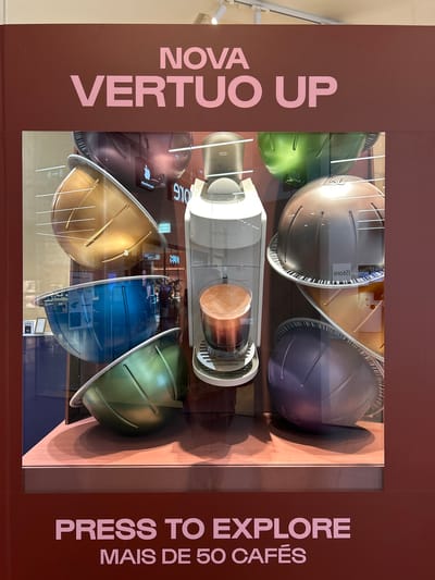 The image shows a display window with a Nespresso Vertuo coffee machine and an assortment of colorful coffee capsules. The text on the display reads "NOVA VERTUO UP" and "PRESS TO EXPLORE MAIS DE 50 CAFÉS." The display is likely in a commercial establishment within Senhora da Hora, Portugal, given the Portuguese text on the display. The scene appears to be a product display, and there are no people visible. The environment is clean and well-lit, suggesting a retail setting. The colors of the capsules are varied, including gold, blue, green, brown, and purple, creating a visually appealing arrangement around the coffee machine.