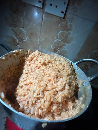 This image captures a large, silver-colored metal pot brimming with reddish-orange cooked rice, consistent with jollof rice, set within an indoor environment in Egbe, Nigeria. The rice appears thoroughly cooked, with individual grains visible along with hints of seasonings or vegetables. The pot, full to its rim, shows one metallic handle on the right side. The background consists of a tiled wall featuring a light brown base with a repeating pattern of white or light-colored orchid-like flowers. Two electrical outlets or switches are visible on the upper part of the tiled wall. No individuals are present in the scene, and no specific actions are depicted beyond the static presentation of the cooked meal.