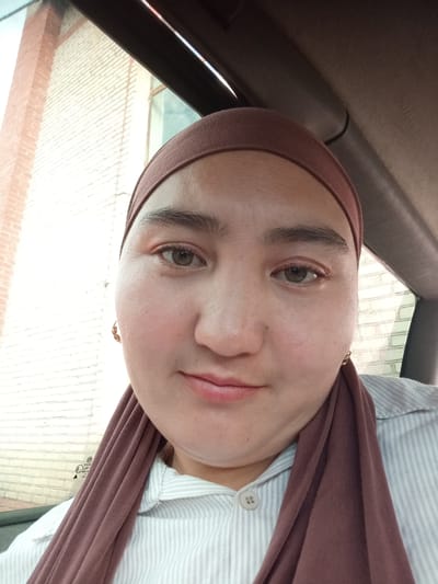 A medium shot, taken from a slightly high angle, shows a young woman wearing a brown hijab and a light-colored striped shirt. She is inside a vehicle, with the interior of the car visible in the foreground. The background outside the car window shows a brick building with a light-colored facade. The woman has brown eyes, lightly applied makeup, and is looking directly at the camera with a slight smile. A small, round gold earring is visible on her left earlobe. The overall lighting suggests it is daytime. The image is assumed to be taken in the city of Kerben, Kyrgyzstan, based on the provided context.