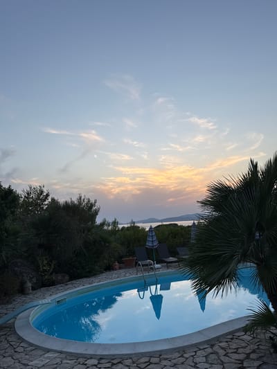 A wide-angle, eye-level shot captures an outdoor swimming pool at dusk. The pool, with a stone border and surrounding paved area, is oval-shaped and filled with clear blue water. The water reflects the sky and surrounding vegetation. Three lounge chairs with umbrellas are positioned by the pool. Lush green foliage and a palm tree frame the scene on the right. In the background, a body of water separates the foreground from distant hills and islands. The sky is a blend of pale blue and soft orange hues, with wispy clouds indicating the setting sun. The location is in Alghero, Italy.