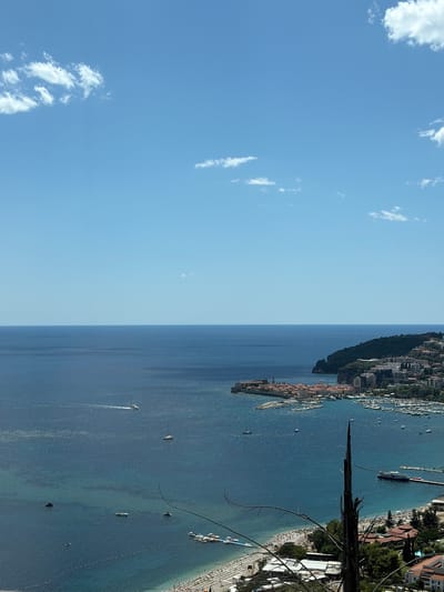 This is a high-angle, medium shot of a coastal city and its harbor on a bright, sunny day. The sky is a clear blue with a few scattered white clouds. The sea is a deep blue, calm, and reflects the sunlight, creating a shimmering effect.

In the foreground, a section of a beach is visible with numerous lounge chairs and umbrellas set up. People are sparsely scattered across the sand. A few boats are moored near the shore.

The middle ground features a picturesque old town with terracotta roofs, situated on a peninsula jutting into the sea. This is identified as the city of Košljun, Montenegro. A harbor is packed with sailboats and smaller vessels. The coastline is lined with buildings, some modern and some older, and a hill covered in dense green foliage rises behind the town.

In the background, the deep blue of the Adriatic Sea stretches to the horizon, meeting the clear blue sky. Several boats, including a larger vessel resembling a ferry or a tour boat, are visible on the water. A rocky outcrop with some vegetation is visible on the far right of the coastline. The overall scene depicts a vibrant coastal town and its active harbor under a serene, sunny sky.