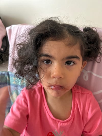 The image shows a close-up portrait of a young girl with dark, curly hair styled in two pigtails. She is wearing a bright pink t-shirt. Her face is partially smeared with a dark substance around her mouth, suggesting she may have recently eaten something messy. Her eyes are open and looking directly at the camera. The background appears to be indoors, with a pink pillow and a patterned blanket visible, suggesting a domestic setting. The lighting is soft and natural, illuminating the girl's face. The image lacks any discernible connection to Boston, United Kingdom.