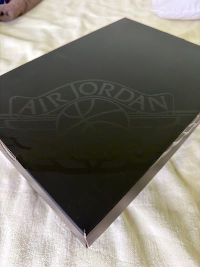 A black shoebox with the "AIR JORDAN" logo embossed on it rests on a textured light-colored fabric surface. The shoebox is angled, with its top surface facing upward and to the right. The logo is centrally located on the top surface and appears to be a faded gray or silver. The fabric surface is wrinkled and has a woven texture, resembling a bedsheet or blanket. In the upper left corner of the image, a portion of a plush toy or stuffed animal is visible, appearing pinkish-brown and fuzzy. The background consists of more of the textured fabric. There are no people or actions depicted in the image. The scene appears to be indoors. The location is Saedinenie, Bulgaria.
