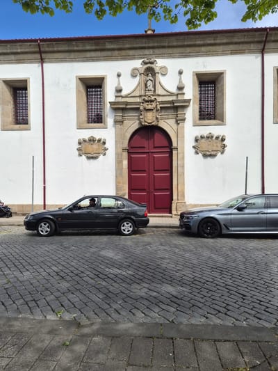 The image shows a white building in Barcelos, Portugal, with a red arched door, barred windows, and stone carvings. Two cars are parked on the cobblestone street in front of the building: a silver car and a black car with a person visible in the driver's seat. Part of a tree is visible in the upper left of the frame.