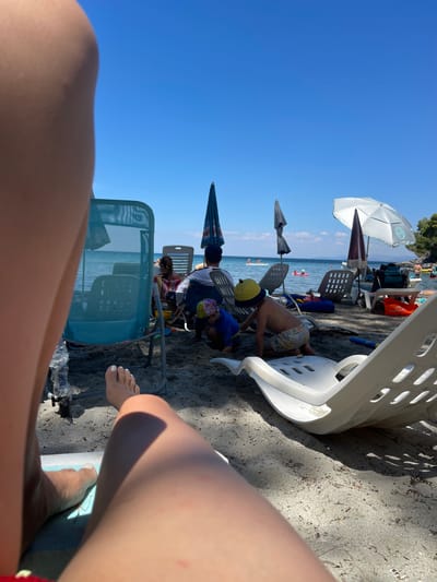 The image depicts a beach scene in Sithonia Municipal Unit, Greece, under a clear blue sky. In the foreground, the legs and feet of a person are visible, suggesting the viewpoint of someone lounging on the sand. Beach chairs, some white and modern, others older, are scattered across the sandy shore. Several beach umbrellas are set up, providing shade.
Further back, people are engaged in various beach activities. Two children are playing in the sand, one in a yellow hat and patterned shorts. Adults are seated in chairs, some observing the children, others looking towards the sea. The shallow water of the Aegean Sea is visible, with a few people swimming or enjoying water activities like paddleboarding. The background shows a tree-lined coastline, indicating a natural, coastal environment. The overall atmosphere is one of a relaxed summer day at the beach.