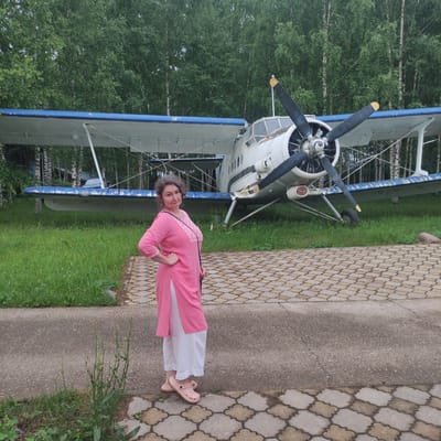 An adult woman of East Asian descent, with curly gray hair and a medium build, stands to the left of a biplane. She is wearing a pink tunic with white trim and white pants, and pink shoes. Her left hand is on her hip, and she is looking towards the camera with a neutral expression. The biplane, which is white with blue accents, has two propellers and is parked on a grassy area. The background is a dense forest of birch trees. The ground in front of the woman and the plane is paved with interlocking decorative stones. The scene appears to be outdoors, possibly in a park or museum, with evidence of a natural environment and a paved pathway.