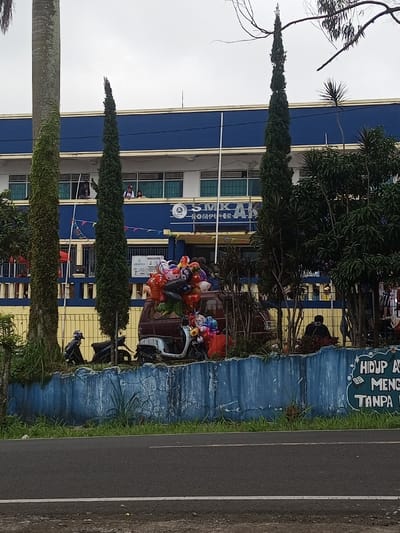 A two-story blue and yellow building with the words "SMK AB" on it is seen behind a row of tall trees and a low, blue wall. A brown vehicle laden with colorful balloons is parked in front of the building. Two motorcycles are parked nearby, and a person is seated on the ground. A street runs in front of the wall. The sky is overcast.