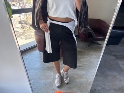 The mirror selfie captures a person indoors, showcasing a casual outfit. The subject wears a white cropped t-shirt, black wide-leg cropped pants, and a white long-sleeved shirt tied around their waist. A dark brown leather-like jacket is draped over their shoulders. Silver metallic Nike sneakers are worn on the feet. Accessories include multiple rings on the fingers, bracelets (one beaded, one with a watch-like element), and white painted fingernails.

The indoor setting features a tiled or polished concrete floor and two armchairs, one brown and one dark. Through a large window, a bright, sunny outdoor scene is visible, depicting a street with utility poles, electrical wires, some vehicles (including a faint white car), and buildings in the distance. The ground outside appears dry and somewhat barren, with a small, light blue object lying on it. The overall impression is daytime, with clear weather in Zovuni, Armenia.