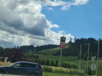 The image shows a road scene with a dark colored SUV in the foreground. In the background, a McDonald's sign is visible, indicating "ARENA FOHNSDORF" and a direction with "5 km". The sign is placed on a pole in front of a hillside covered with green grass and trees. The sky is blue with scattered white clouds. The overall environment appears to be rural or suburban, with indications of a roadside location. Based on the provided information, this scene is located in or near Judenburg, Austria.