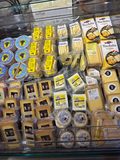 The image depicts a display of various Swiss cheeses in a refrigerated display case, likely in a grocery store or a cheese shop. The cheeses are packaged in plastic containers and wrappers. Prominently visible are "Raclette Classique" packages, "Urner Alpkäse," "Urner Ziegenkäse," "Sittisalp," and "Rahm-Chäsli." The labeling indicates that these are products from Switzerland. Some labels include images of cows and scenic mountain landscapes. A sign overhead reads "Aktion," suggesting a special offer or promotion. The scene appears to be indoors, with artificial lighting illuminating the products. The general appearance suggests a clean and organized retail environment. Although the image itself does not explicitly state the location as Schattdorf, Switzerland, the presence of "Urner" products, a canton in Switzerland, strongly suggests a location within that region, and Schattdorf is a municipality in the canton of Uri, Switzerland, which is home to Urner Alpkäse and other Urner products.