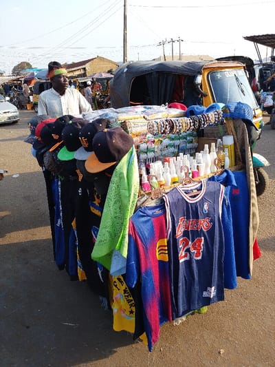 A vendor stands behind a three-wheeled vehicle overflowing with goods. Hats, clothing, jewelry, and small bottles are displayed. A Lakers basketball jersey and a Barcelona soccer jersey hang at the front. The vendor wears a white shirt and a colorful hat. Other people and vehicles are visible in the background, suggesting a busy market area. The sky is bright and clear.