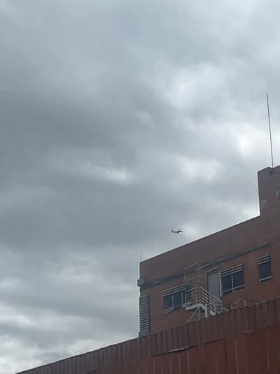 The image displays an urban outdoor scene in Bogota, Capital District, Colombia, characterized by an overcast sky. The upper two-thirds of the frame are filled with dense, uniform grey clouds. In the upper-mid-right quadrant, a small, dark silhouette of an airplane is visible in flight against the cloudy background. The lower right portion of the image is occupied by a multi-story building constructed of red brick. This building features multiple rectangular windows with dark frames and an external metal staircase with white railings leading up to a door on its visible façade. A slender metal rod, consistent with a lightning rod or antenna, extends vertically from the building's flat roof on the right side. A lower red brick wall or structure is present in the foreground, extending across the bottom of the frame and partially obscuring the base of the main building. No individuals are present in the depicted scene.