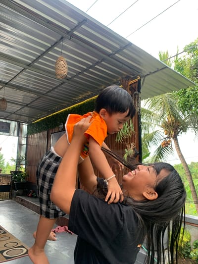 In Sukasada, Indonesia, a young woman with long, dark hair is holding a small child aloft, with the child looking down at her. The woman is wearing a black t-shirt, and the child is wearing an orange t-shirt and black and white checkered shorts. They are outdoors under a metal roof structure, which appears to be part of a home or restaurant. The background shows a bamboo wall, green foliage, and a coconut tree. The lighting suggests it is daytime, possibly overcast.