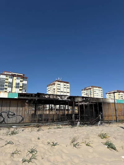 In Costa da Caparica, Portugal, a burnt-out structure sits on a sandy beach under a clear blue sky. The building, likely a beachside establishment, shows significant fire damage, with charred remains of its roof and walls visible. The exterior of the structure is lined with vertical corrugated metal panels, some of which are rusted and discolored. Graffiti is present on some of these panels. Behind the damaged building, four multi-story apartment buildings stand tall, their facades a mix of white, yellow, and brown. Greenery, including sparse beach grass, grows in the sand in the foreground. The scene conveys a sense of destruction and abandonment, contrasting with the orderly apartment buildings in the background.