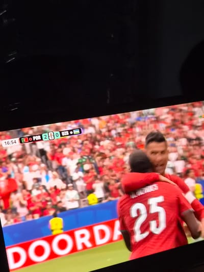 This media file captures a close-up view of a television screen displaying a football (soccer) match. The screen shows a player in a red jersey with the number "25" prominently displayed on the back. The player's face is partially visible, looking down. The background is filled with a crowd of spectators wearing red, suggesting they are fans of one of the teams.
Along the bottom edge of the screen, a scoreboard graphic is visible, indicating the match is between POR and UZB, with the score being 2-0. The time displayed is 16:54. Another visible graphic reads "DOORDA" vertically in large white letters on a red background, likely an advertisement.
The image appears to be taken in a dark room, with the television screen being the primary light source. The overall scene suggests a live sporting event being broadcast. The setting appears to be a living room or similar indoor space where a television is being watched. The image is slightly blurred and the colors are somewhat oversaturated, common characteristics of a photograph taken of a screen. The provided location context of Differdange, Luxembourg, indicates where this viewing may be taking place.