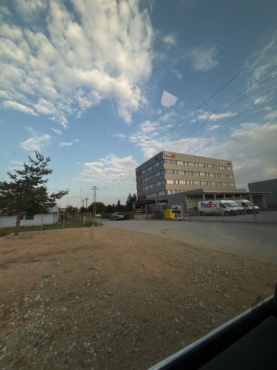 The image displays an outdoor scene captured from inside a vehicle, looking towards a commercial property in Tsaratsovo, Bulgaria.
The sky occupies the upper portion of the frame, showing a clear blue sky with scattered white cumulus clouds. Power lines stretch horizontally across the upper-mid section of the sky. A distinct, bright, rectangular reflection is visible on the vehicle's window in the upper-center of the sky.
The foreground consists of an unpaved, gravelly, and dirt-covered area, indicating an undeveloped or construction zone. The bottom right corner reveals the dark interior trim and glass edge of the vehicle's window.
On the left side of the midground, a solitary, medium-sized evergreen tree, likely a pine, stands on the dirt ground. Behind it, a small, light-colored, single-story building with a dark roof is partially visible, featuring at least one window. A light-colored fence with metal posts extends from the left towards the center. Further back, several utility poles with visible wires are present, along with more distant trees. A dark-colored SUV is parked on an asphalt road in the mid-distance on the left.
The dominant feature on the right side of the midground is a multi-story, rectangular commercial building, constructed with a grey/silver facade and numerous uniformly spaced windows across its multiple floors. Prominent red "FedEx" logos are affixed to the top of the building, including one on the front-facing corner and another centrally placed on the visible side. Additional logos, "AlphaSat" and "TEB," are also visible near the top of the building. Below the main structure, a grey covered loading dock or service entrance is present. Inside a fenced area immediately in front of this dock, three white "FedEx" delivery vans are parked. A metal chain-link fence separates this operational area from the dirt foreground. Near the fence line, a yellow dumpster or set of bins is visible next to a larger grey industrial waste bin.
The background beyond the main building shows more distant trees and open sky, suggesting a semi-rural or industrial park setting. The lighting suggests daytime conditions, possibly late afternoon given the softer light and absence of harsh shadows. No individuals are clearly visible in the scene.